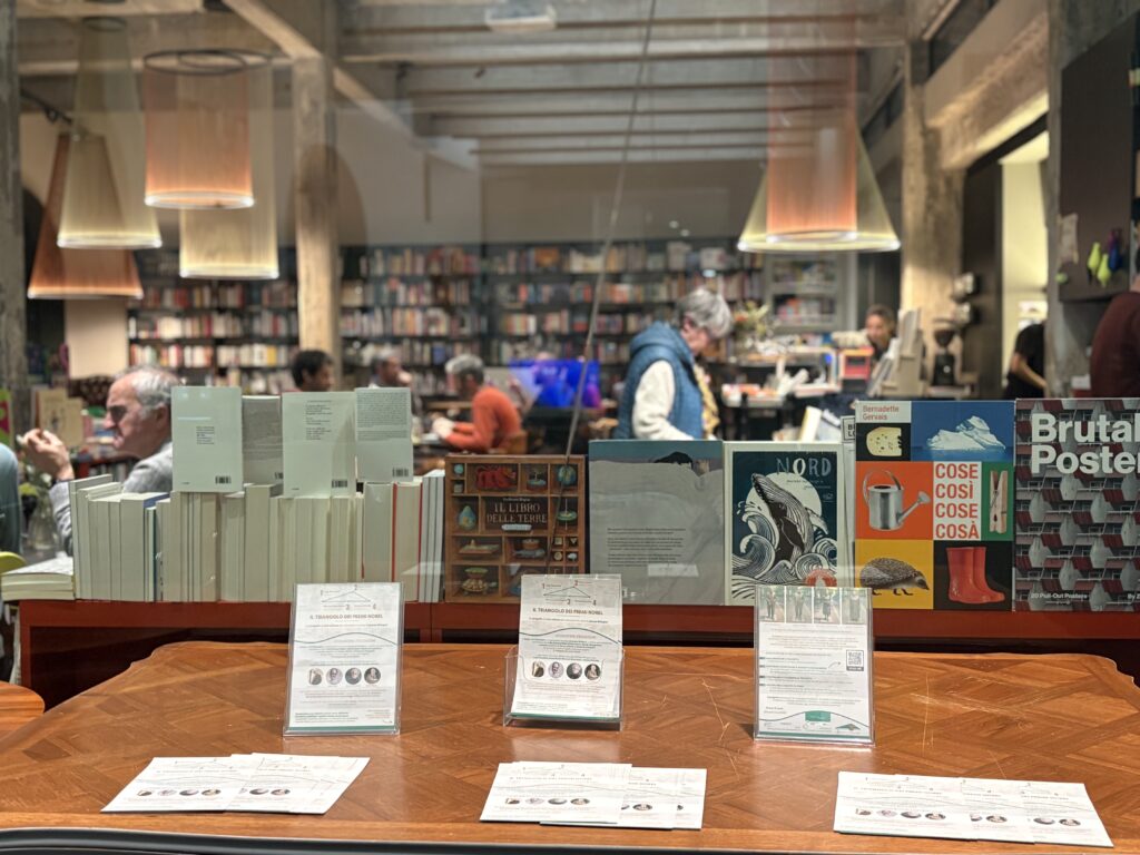 Foto scattata ad un nostro evento da Shell Libreria Bistrot, in primo piano i volantini del Progetto, sullo sfondo la bellissima cornice di Shell, con sagome di persone intente a leggere o a conversare.